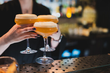 A bartender holds three vibrant orange-yellow, foam-topped passion fruit martinis in coupe glasses over a bar counter, ready to serve in a dimly lit bar setting
