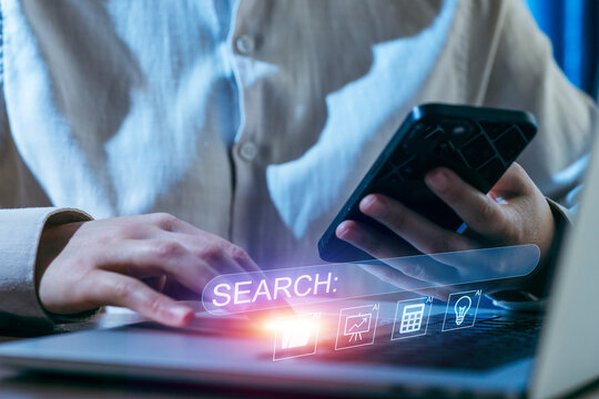 Person using a smartphone to conduct online research while working on a laptop in a dimly lit room