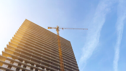 Obraz premium Highrise building under construction with a tall yellow tower crane lifting materials, illuminated by warm sunlight against a clear blue sky, symbolizing urban development and modern architecture