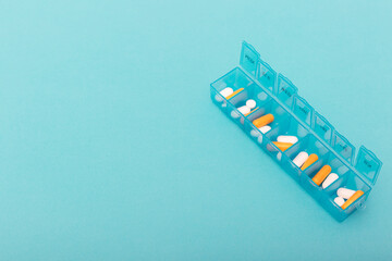 Blue plastic pill organizer with orange and white capsules on turquoise background. Copy space