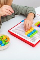 Child arranges colorful plastic pegs on a red mosaic board. The photo illustrates fine motor skill development, early learning, focus, and creativity through educational play. Vertical photo