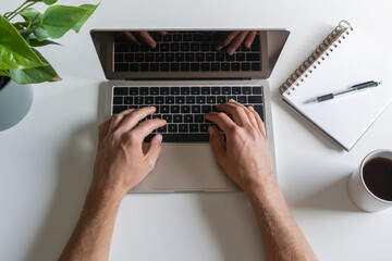Male hands on laptop on white table, female with coffee or tea, notepad and pen nearby