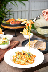 A hand lifting chicken spaghetti coated in rich tomato sauce with a fork, captured with softly blurred seafood dishes in the background, creating a warm and appetizing dining-scene presentation.