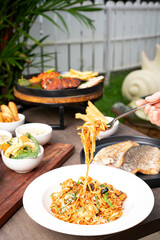 A hand lifting chicken spaghetti coated in rich tomato sauce with a fork, captured with softly blurred seafood dishes in the background, creating a warm and appetizing dining-scene presentation.