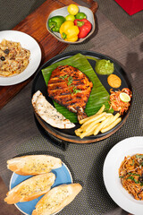 Top shot of tandoori pomfret masala served with chutney, salad, fries, and kulcha on a plate, styled alongside chicken spaghetti, cheesy penne seafood, garlic bread, and raw capsicums 