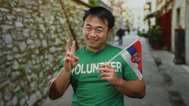 Young man wearing green shirt holding serbian flag in urban street, symbolizing cultural pride and volunteer spirit outdoors.