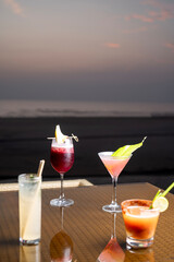 Elegant shot of four colorful cocktails and their reflections on a glass table with the calm sea and vibrant post-sunset sky in the background, creating a stylish tropical evening ambience.