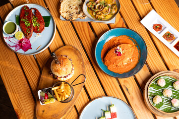 Top shot of a food spread on a brown table with Goan fish curry rice, kingfish rechado, chicken burger with fries, butter chicken naan, truffle dim sum, and watermelon feta salad.