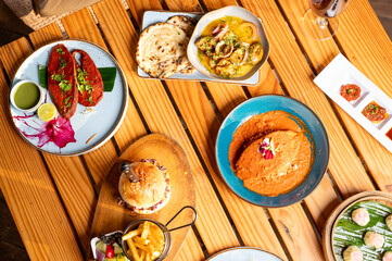 Top shot of a food spread on a brown table with a Goan fish curry rice, kingfish rechado masala, double chicken burger with fries, butter chicken naan, and truffle edamame dim sum.
