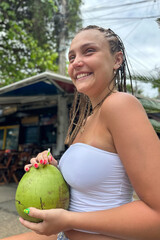 Young woman holding a fresh green coconut in front of lush tropical greenery. Vibrant summer atmosphere, travel lifestyle, exotic vacation mood, nature background.