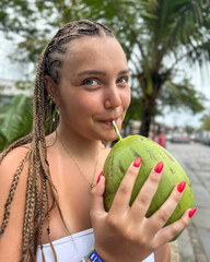 Young woman holding a fresh green coconut in front of lush tropical greenery. Vibrant summer atmosphere, travel lifestyle, exotic vacation mood, nature background.
