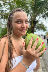 Young woman holding a fresh green coconut in front of lush tropical greenery. Vibrant summer atmosphere, travel lifestyle, exotic vacation mood, nature background.