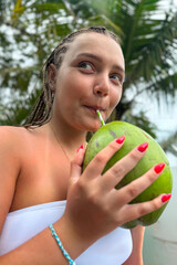 Young woman holding a fresh green coconut in front of lush tropical greenery. Vibrant summer atmosphere, travel lifestyle, exotic vacation mood, nature background.