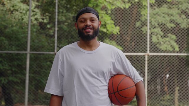 Smiling African American man wearing durag holding basketball outdoors, standing in court with friendly confident expression, relaxed pose and natural demeanor