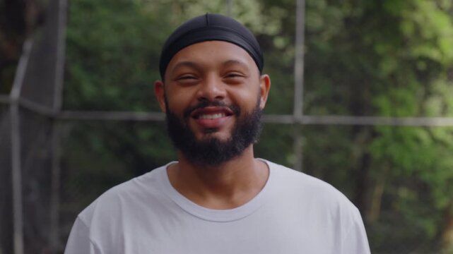 Smiling African American man wearing durag standing outdoors in basketball court, looking directly at camera with warm friendly expression and relaxed confident demeanor