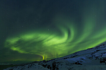 Spectacular wide arc of intense green Aurora Borealis soaring high above a snow-covered rocky ridge...