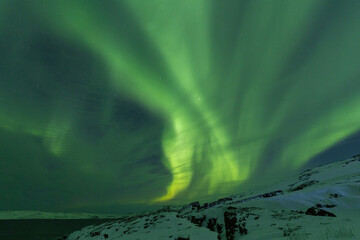 Spectacular wide arc of intense green Aurora Borealis soaring high above a snow-covered rocky ridge...