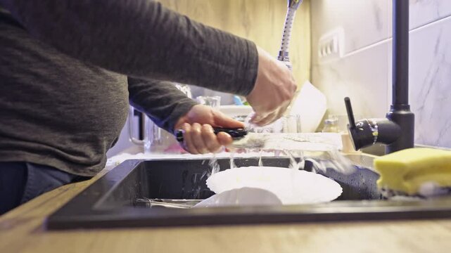 A person diligently washes and rinses dishes under running water in a contemporary kitchen sink, highlighting the routine of domestic cleanliness and home maintenance.