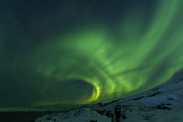 Spectacular wide arc of intense green Aurora Borealis soaring high above a snow-covered rocky ridge...