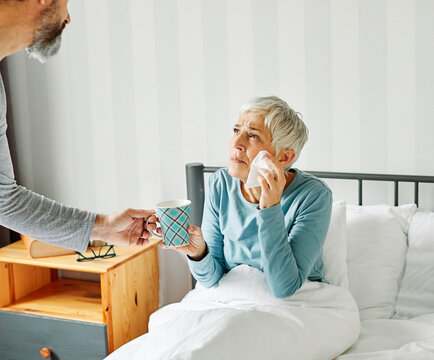 Portrait of a sick senior elderly woman sneezing and using tisue in bed in the morning, a mature woman at home, cold or flu illness and pain, senior couple, husband offering hot tea to his wife, love 
