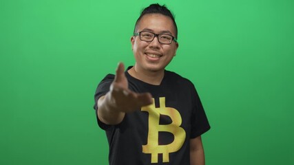 Man wearing bitcoin t shirt extends hand toward camera and smiles in green studio; trust invitation.