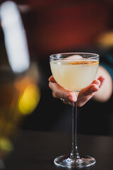 A person is holding a light-colored cocktail in a coupe glass, garnished with an orange or lime slice, in a dimly lit bar setting. The focus is sharp on the glass and hand