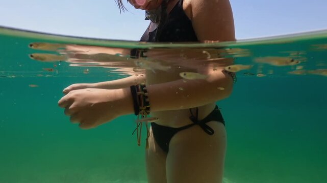 Young woman in black bikini stands in crystal-clear turquoise lake with snorkel mask, feeding small curious fish that swim playfully around her outstretched hands on a sunny summer day