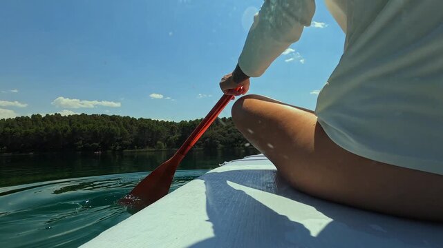Millennial woman sitting on stand up paddle board, paddling in slow motion on beautiful lake, enjoy sunny day outdoors