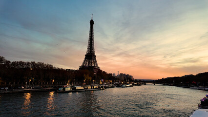 Fototapeta premium A dramatic, high-contrast twilight view of the Eiffel Tower silhouetted against a blue-orange gradient sky, emphasizing the shimmering water reflections of the barges on the Seine.