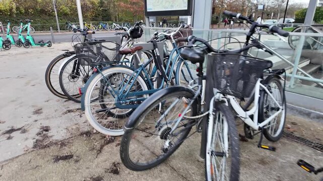 A dynamic close-up of commuter bicycles packed tightly in a modern rack, featuring muted autumn light and a glass public transport escalator in the background.