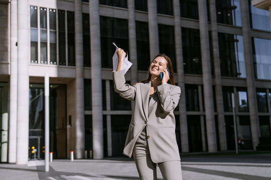 A joyful businesswoman celebrates while chatting on the phone, waving a paper, with tall office buildings in the background.