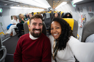 Happy couple taking a selfie inside an airplane cabin during flight. Smiling man and woman in casual clothes with rows of seats and passengers, wide angle view conveys travel joy and vacation vibes