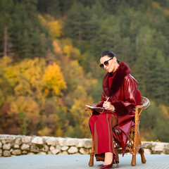 Fashion director in a burgundy leather coat sitting in a wicker chair and writing notes during an outdoor autumn photoshoot. Confident creative leader planning the next scene with focus and elegance.