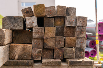 Stack of square wood with rusty nails, next to a stack of plastic pipes on a construction site.