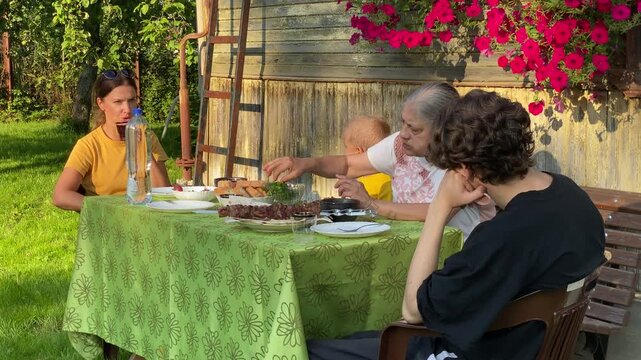 A three-generation family&mdash;grandmother, mother, and two children&mdash;sits around an outdoor table, enjoying a delicious barbecue meal. This summer dinner is taking place outside an old, rustic wooden house