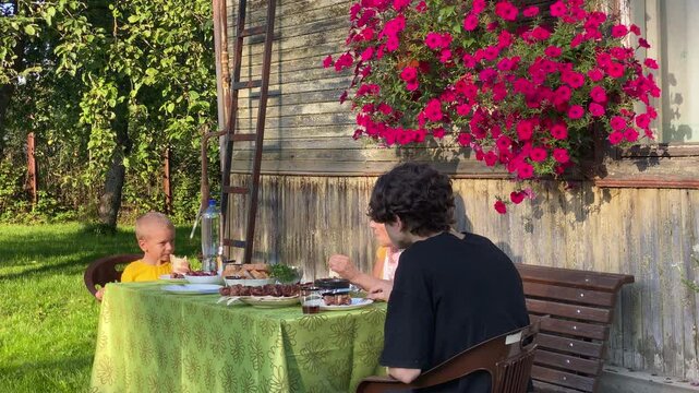 A three-generation family&mdash;grandmother, mother, and two children&mdash;sits around an outdoor table, enjoying a delicious barbecue meal. This summer dinner is taking place outside an old, rustic wooden house