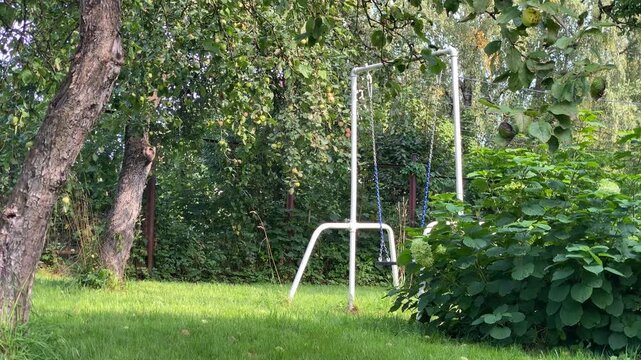 A simple white metal swing set stands on a bright green lawn, surrounded by lush trees and bushes, including an apple tree with fruit. This peaceful backyard scene captures the natural beauty