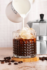 Cream Milk is pouring into Iced brew coffee. Cold Coffee drink cocktail with ice and moka pot on beige table background. Summer refreshing drink