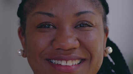 Smiling African American woman looking directly at camera with warm confident expression, wearing pearl earrings and braided hair in softly lit indoor setting
