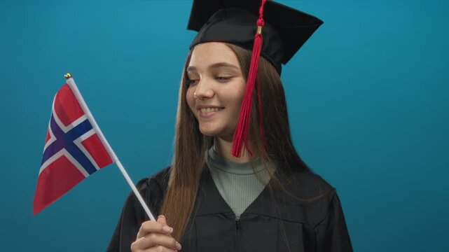 Teenage girl wearing cap and gown holds norwegian flag with smiling face in blue studio; pride accomplishment future success.