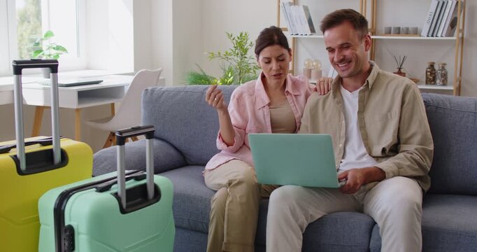 Discussion of happy couple planning travel and summer vacation together, people sitting on couch by suitcases at home. Young man and woman discuss online hotel and flight booking choice, using laptop