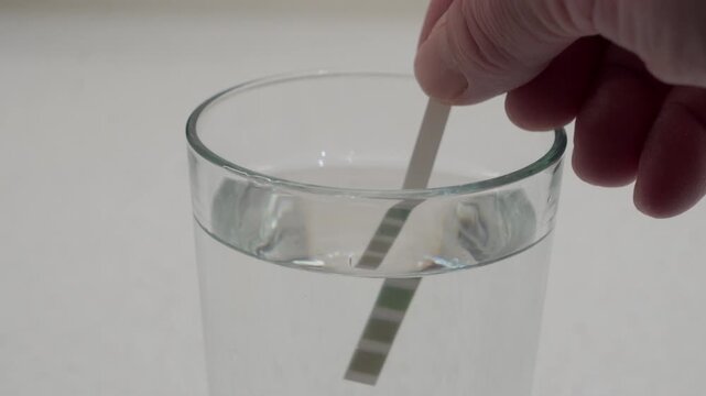 A hand dips a water hardness testing strip into a glass of clear water. The test is performed to determine the mineral content and quality of the water supply.
