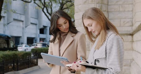 Two professionals review documents in urban setting, Pair of businesswomen collaborating on project near city building - Powered by Adobe