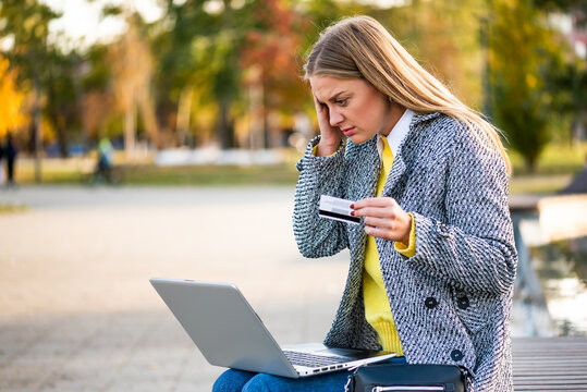 Businesswoman in autumn coat looking stressed while using laptop and holding credit card. She is dealing with financial problems while working remotely in the city park. - Powered by Adobe