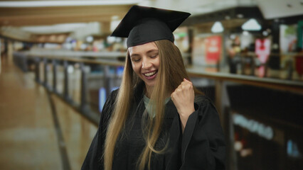 Young woman in graduation attire smiles joyfully in a busy shopping mall, embodying success and happiness amidst store signs and bustling atmosphere indoors.