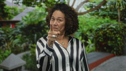 Woman wearing striped shirt shows thumbs up gesture in building with green foliage; optimism hope...