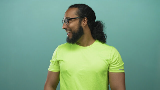 Man with beard and glasses smiling and facing sideways in studio with teal background wearing neon green shirt and curly ponytail; happiness welcome. - Powered by Adobe