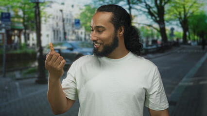 Man holding cookie with hand on amsterdam canal street, smiling and examining the biscuit; casual happiness.