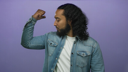 Man flexes bicep in studio purple backdrop wearing denim jacket and white t shirt with fist raised to shoulder; confidence strength.