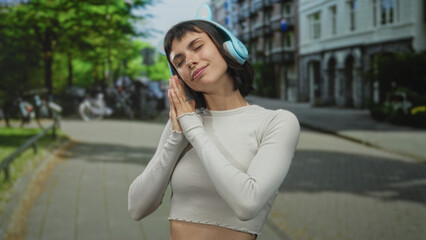 Young hispanic woman presses palms together wearing blue cat ear headphones on street with building and trees; serenity.